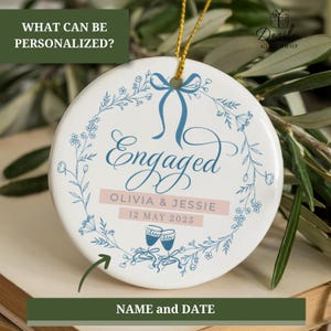 May include: A round white ceramic ornament with the word "Engaged" in blue script, encircled by a floral wreath. It features the names "Olivia & Jessie" and the date "12 MAY 2025". A gold string is attached for hanging.