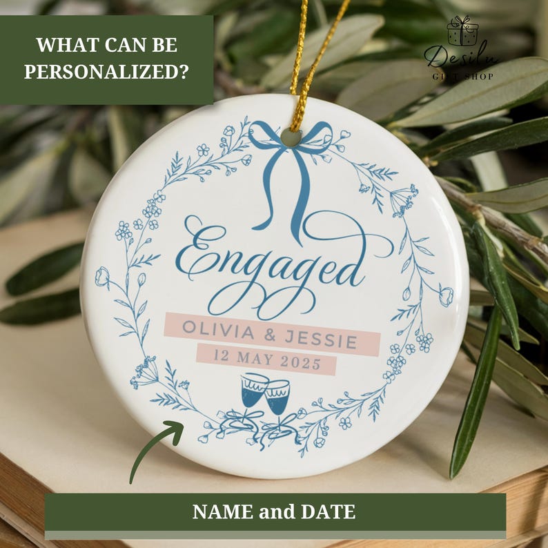 May include: A round white ceramic ornament with the word "Engaged" in blue script, encircled by a floral wreath. It features the names "Olivia & Jessie" and the date "12 MAY 2025". A gold string is attached for hanging.