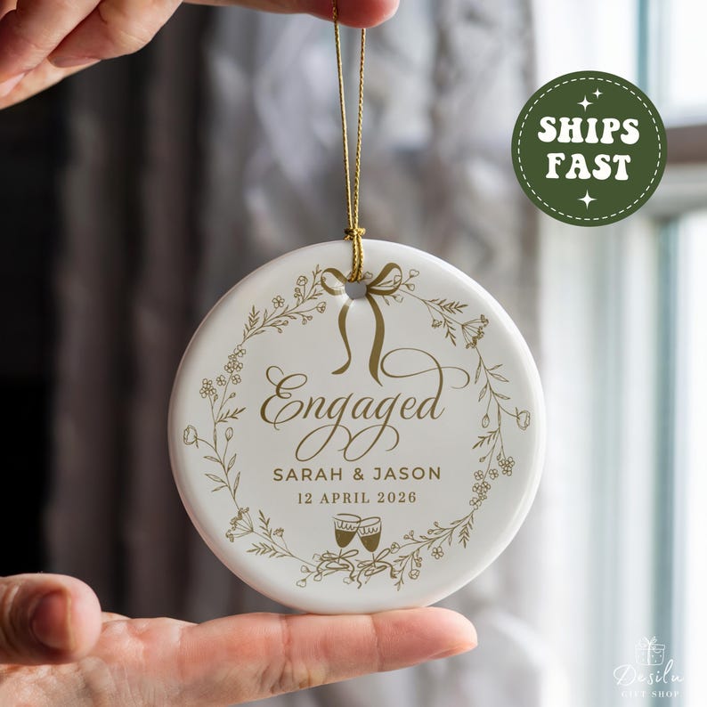 May include: A round, white ceramic engagement ornament with gold text and a gold hanging ribbon. The ornament features the word "Engaged", names, and a date in gold script, surrounded by a floral wreath design. The date is written as 12 April 2026.