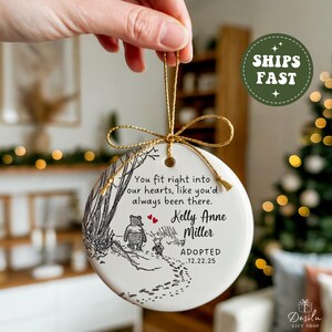 K&ouml;nnte beinhalten: Runder, wei&szlig;er Keramik-Weihnachtsschmuck mit goldener Schnur. Er zeigt eine Schwarz-Wei&szlig;-Illustration von Winnie Puuh und Ferkel. Der Schmuck tr&auml;gt den Namen "Kelly Anne Miller" und das Wort "ADOPTED".