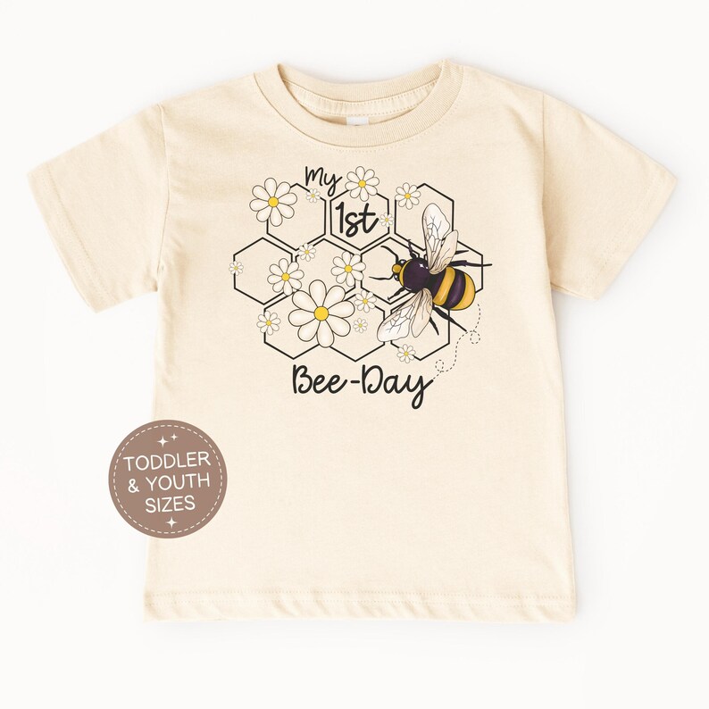 May include: Cream-colored toddler t-shirt with a bee-themed graphic. The design features a bee, daisies, and honeycomb shapes, with the text "My 1st Bee-Day." The shirt is available in toddler and youth sizes.