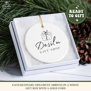K&ouml;nnte beinhalten: Wei&szlig;er Keramikschmuck mit goldener Kordel, in einer wei&szlig;en Geschenkbox. Der Schmuck zeigt eine schwarze Strichzeichnung eines Geschenks und den Text "Desilu GIFT SHOP". Die Box ist von Tannennadeln und Tannenzapfen umgeben. Der Text "READY TO GIFT" ist ebenfalls sichtbar.