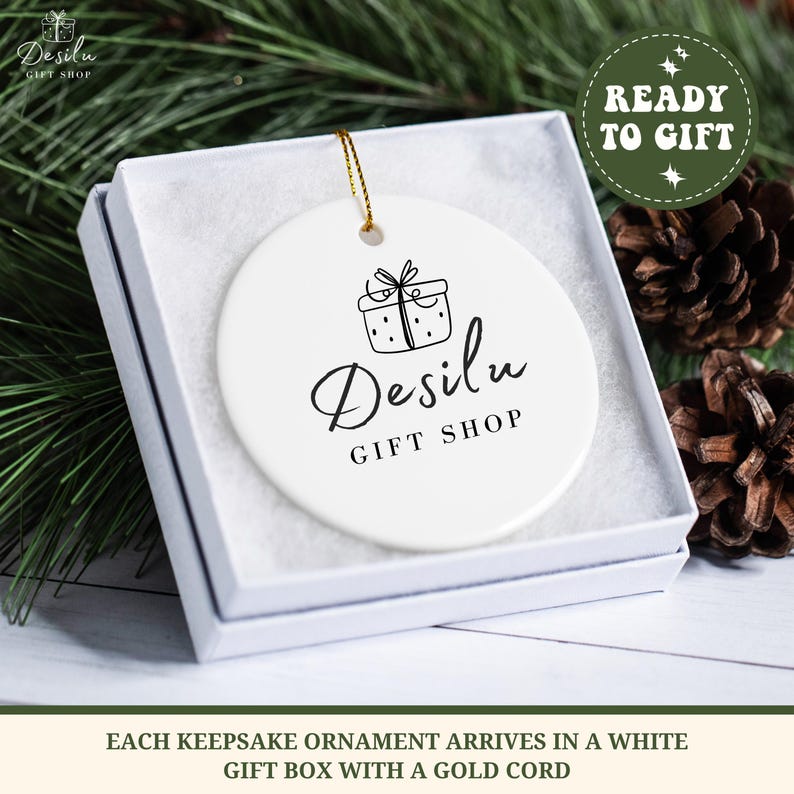 K&ouml;nnte beinhalten: Wei&szlig;er Keramikschmuck mit goldener Kordel, in einer wei&szlig;en Geschenkbox. Der Schmuck zeigt eine schwarze Strichzeichnung eines Geschenks und den Text "Desilu GIFT SHOP". Die Box ist von Tannennadeln und Tannenzapfen umgeben. Der Text "READY TO GIFT" ist ebenfalls sichtbar.