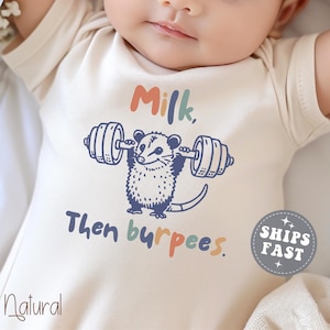 May include: Cream-colored baby onesie with a playful possum graphic lifting weights. Text reads "Milk, Then burpees." The word "Natural" is printed at the bottom left. A circular badge with the words "SHIPS FAST" is on the right.