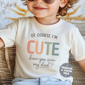 May include: Cream-colored toddler t-shirt with the text "OF COURSE I'M CUTE have you seen my dad?" in colorful letters. The shirt also has a small circular logo that says "TODDLER & YOUTH TEES".