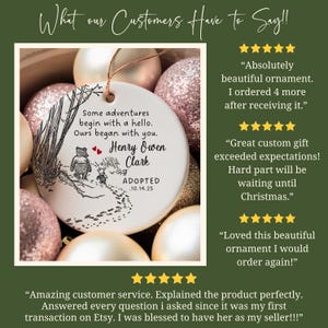 K&ouml;nnte beinhalten: Ein wei&szlig;er Keramik-Weihnachtsschmuck mit einem Winnie-Puuh-Design. Der Schmuck tr&auml;gt den Text "Some adventures begin with a hello. Ours began with you. Henry Owen Clark ADOPTED 10.14.25". Der Schmuck ist von goldenen und rosa Weihnachtskugeln umgeben.