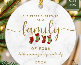 May include: White ceramic Christmas ornament with a marble pattern. It says "Our First Christmas as a Family of Four" with the names "daddy, mommy, alyssa, brody" and the year "2024". There are four red and green stockings hanging from the top.