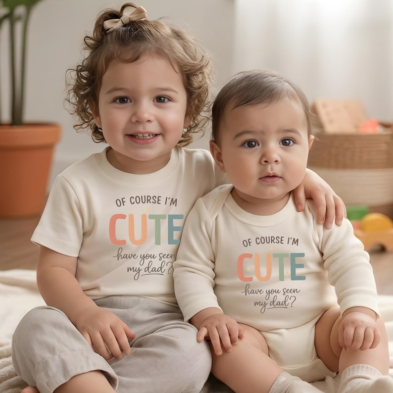May include: Two children wearing matching cream-colored outfits with the text "OF COURSE I'M CUTE have you seen my dad?" in colorful letters. One child wears a t-shirt, the other a onesie. Both are sitting on a light-colored blanket.