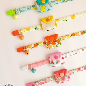 May include: Five fabric collars with attached small pouches, each featuring fruit patterns including lemons, pineapples, oranges, watermelons, and peaches. Each collar includes a plastic clip, a metal ring, and a small bell in matching colors.