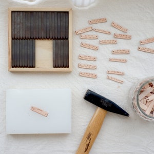 May include: A set of metal letter stamps in a wooden box, a hammer, and several leather tags with names stamped on them. The tags are in a glass jar and on a white surface.