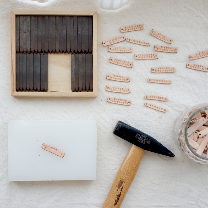 May include: A set of metal letter stamps in a wooden box, a hammer, and several leather tags with names stamped on them. The tags are in a glass jar and on a white surface.