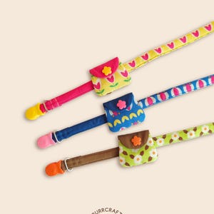 May include: Three fabric pacifier clips with small attached pouches. The top strap is yellow with pink tulips and a yellow flower button. The middle is blue with a pink flower button. The bottom is green with white flowers and an orange button.