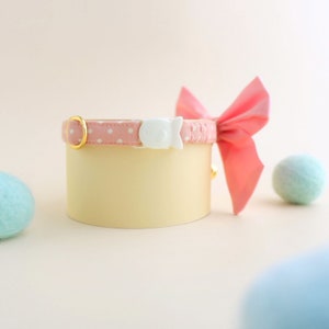 May include: Pink and white polka dot cat collar with a white plastic safety buckle and a pink bow. The collar is on a light brown surface.