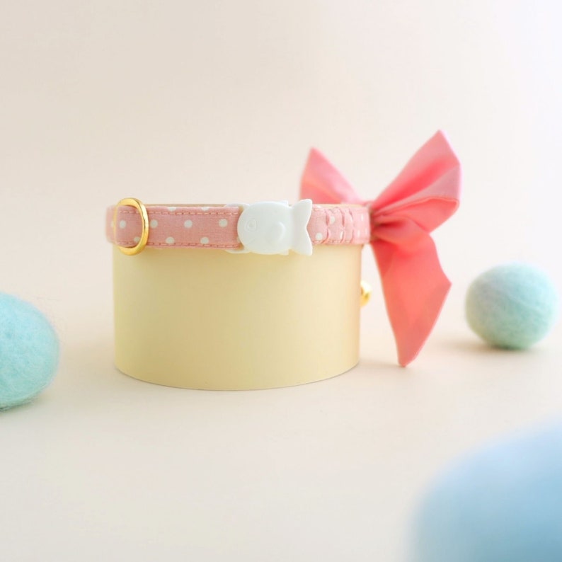 May include: Pink and white polka dot cat collar with a white plastic safety buckle and a pink bow. The collar is on a light brown surface.