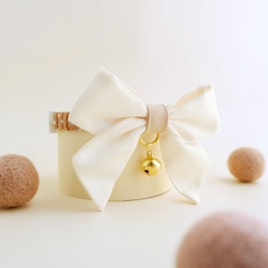 Peut inclure: Un nœud papillon en tissu blanc avec un charme de cloche dorée. Le nœud papillon est attaché à un collier à motif brun et blanc.