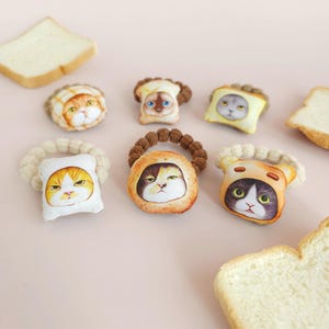 May include: A collection of cat-themed hair ties, each featuring a different cat face design. The ties are round and made of fabric, with the cat faces printed on them. Slices of bread are scattered around the hair ties on a pink surface.