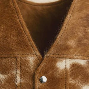 May include: Close-up of a brown and white cowhide vest. The vest features a V-neck design and a single silver snap closure. The stitching is visible, and the fur has a natural pattern.
