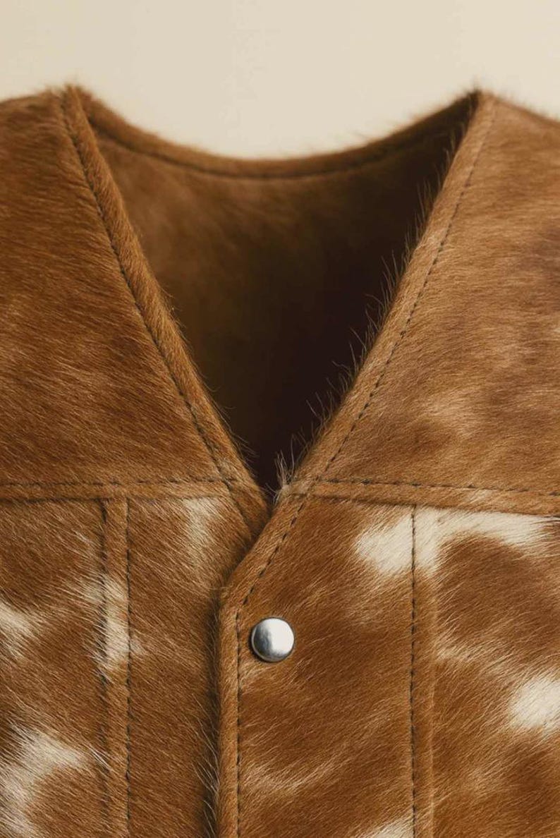 May include: Close-up of a brown and white cowhide vest. The vest features a V-neck design and a single silver snap closure. The stitching is visible, and the fur has a natural pattern.