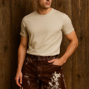 May include: A person wearing a beige crew neck t-shirt and brown leather shorts with white cowhide patches. The shorts have a brown leather belt and pockets. The background is a wooden wall.