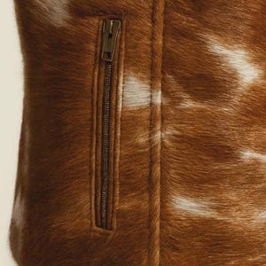 May include: Close-up of a brown and white cowhide jacket with a zipper. The jacket has a natural, textured appearance with a mix of brown and white fur. The zipper is a dark bronze color.