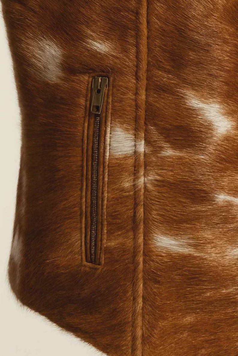 May include: Close-up of a brown and white cowhide jacket with a zipper. The jacket has a natural, textured appearance with a mix of brown and white fur. The zipper is a dark bronze color.