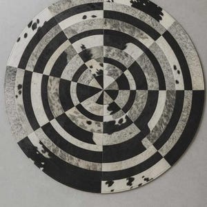 Peut inclure: Tapis rond au design géométrique. Il présente des cercles concentriques et des carrés en noir, blanc et gris. La texture du tapis semble être un mélange de matériaux, avec des sections ressemblant à du cuir animal.