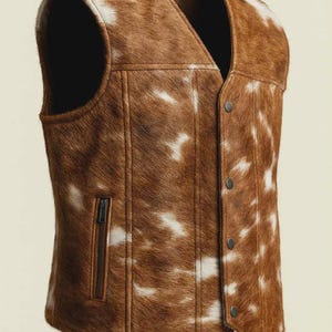 May include: A brown and white cowhide vest with a V-neck and a button-up front. The vest has a zippered pocket on the left side and is made of a soft, textured material. The vest is a medium size.