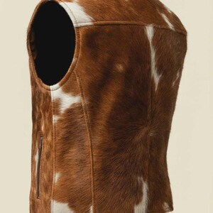 May include: A brown and white cowhide vest with a zipper closure. The vest has a round neckline and a tailored fit. The natural pattern of the cowhide is visible, with brown and white patches. The vest is a unique piece of outerwear.