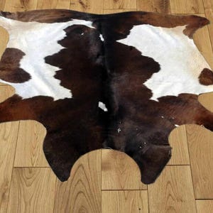 Puede incluir: Una alfombra de piel de vaca con un patrón natural de manchas marrones y blancas. La alfombra tiene una forma irregular, con un centro marrón oscuro y bordes marrón claro. Las manchas blancas están dispersas por la superficie, creando un diseño único.