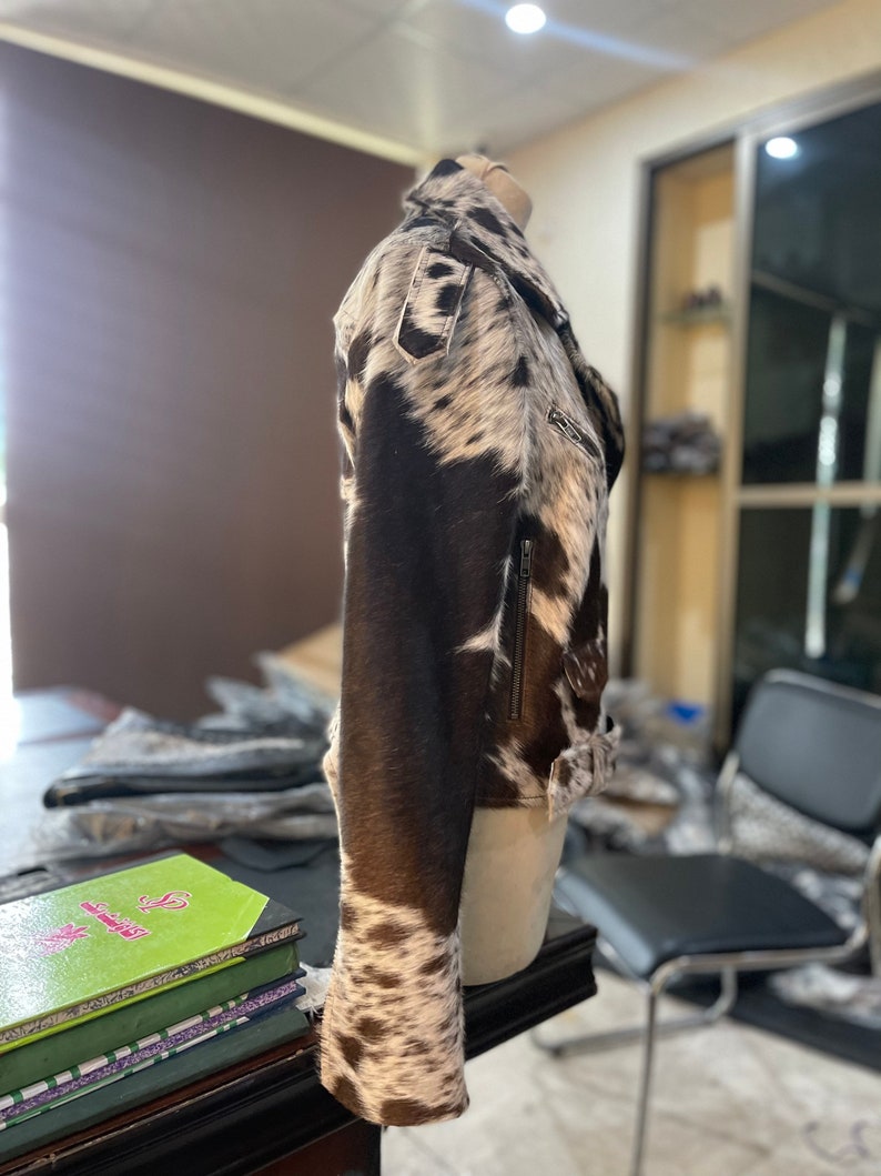 May include: A brown and white cowhide leather jacket with a zipper closure. The jacket has a stand-up collar and long sleeves.