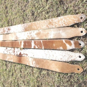May include: Five brown leather guitar straps with white cowhide patches. The straps are laid out in a row on a green grassy surface.