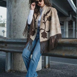 May include: A beige cape with dark brown floral patterns and long fringe trim, worn over a white button-down shirt, blue denim jeans, and dark brown heels. The outfit is accessorized with dark-tinted sunglasses.