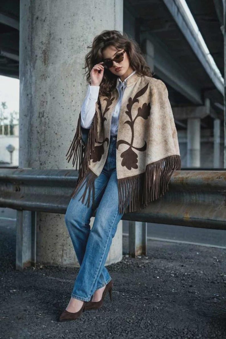 May include: A beige cape with dark brown floral patterns and long fringe trim, worn over a white button-down shirt, blue denim jeans, and dark brown heels. The outfit is accessorized with dark-tinted sunglasses.