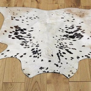 Puede incluir: Alfombra de piel de vaca de forma irregular, principalmente blanca con manchas negras. La alfombra está colocada sobre un suelo de madera, mostrando su patrón natural.