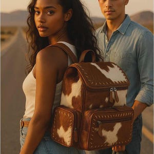 May include: A brown and white cowhide backpack with multiple pockets and a top handle. The backpack has a rectangular shape with a flap closure and is worn by a person. The background shows a desert road.