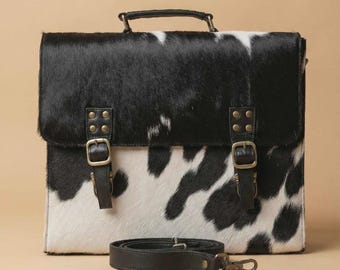 May include: A black and white cowhide briefcase with a top handle and a detachable shoulder strap. The bag features two front straps with brass buckles and a flap closure. The briefcase is rectangular and has a black and white spotted pattern.
