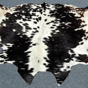 Puede incluir: Una alfombra de piel de vaca con un patrón natural de manchas negras y blancas. La alfombra tiene una forma irregular, con bordes curvos y extensiones puntiagudas. La piel es suave y parece estar bien curtida, adecuada para cubrir el suelo.