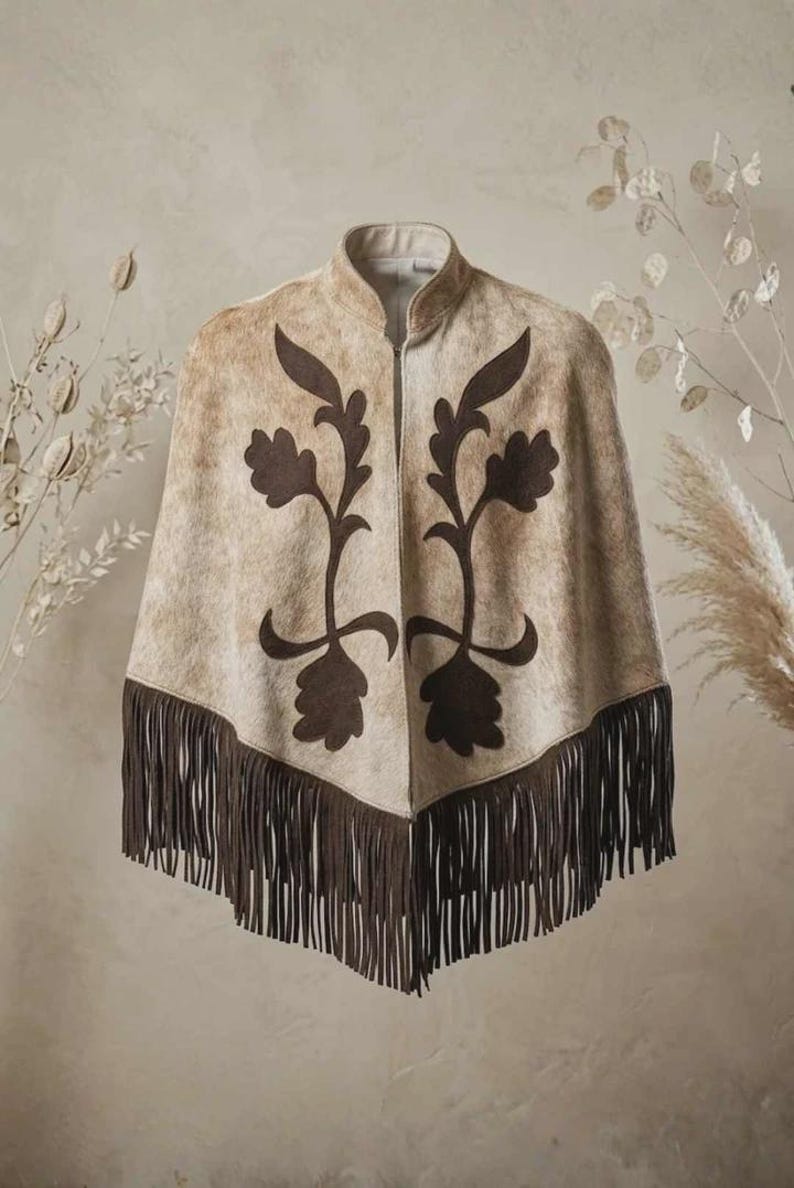 May include: A beige and tan textured poncho featuring a high collar and a front zipper closure. The garment is decorated with dark brown symmetrical floral applique patterns on the front and finished with a long dark brown fringe hemline.