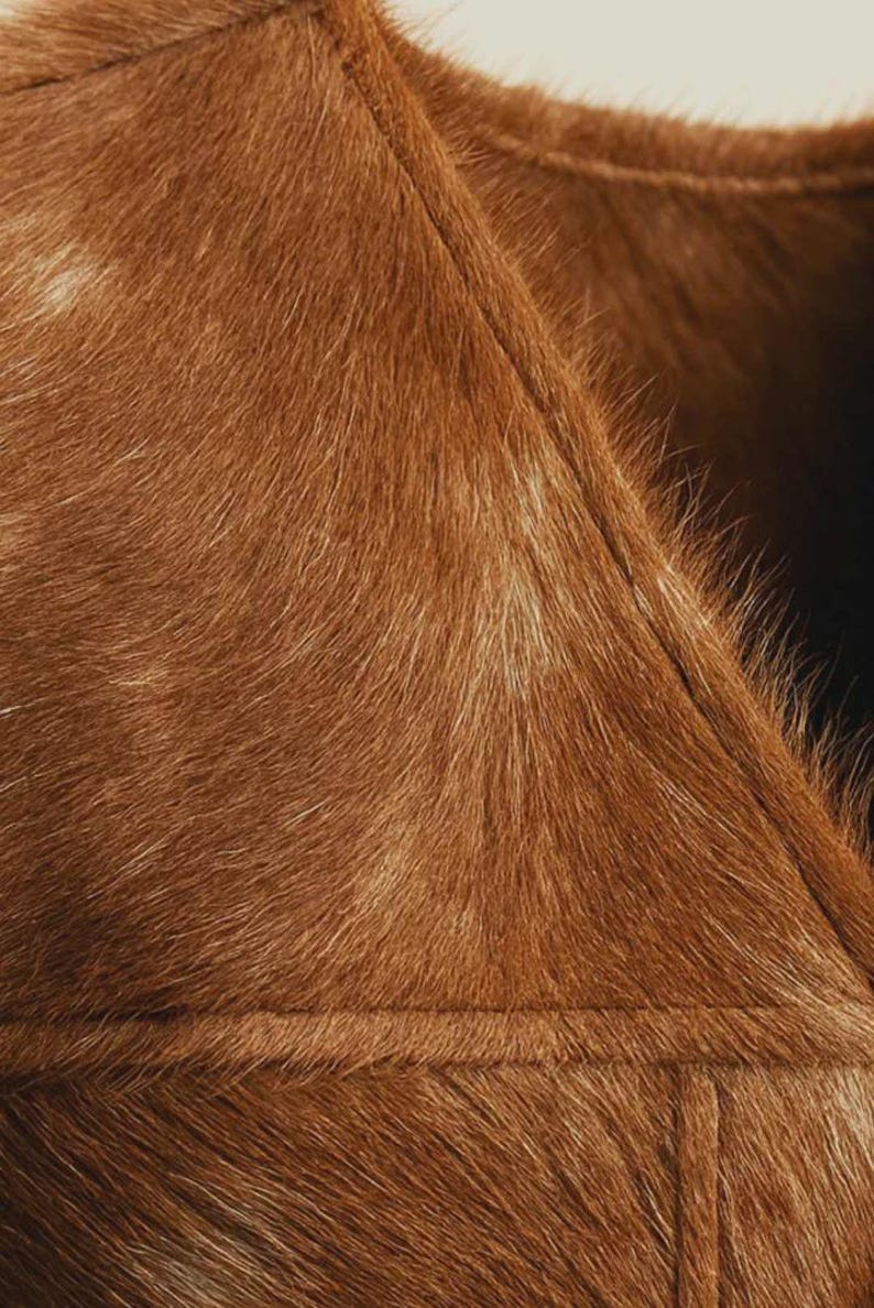 May include: Close-up of a brown fur garment, possibly a vest or coat. The fur is a rich, warm brown with lighter highlights, and the stitching is visible. The texture appears soft and luxurious, suggesting a high-quality material.