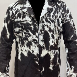 Puede incluir: Una chaqueta larga de piel de vaca negra y blanca con cierre de botones y mangas largas.