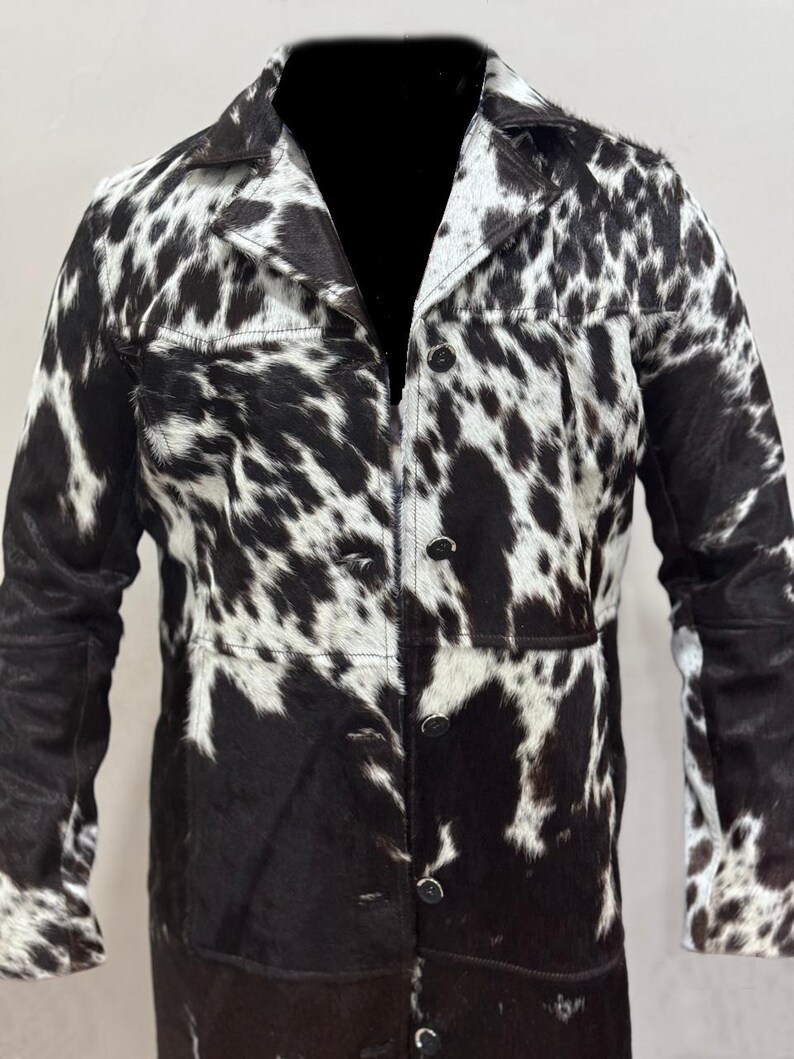 Puede incluir: Una chaqueta larga de piel de vaca negra y blanca con cierre de botones y mangas largas.