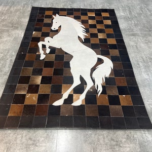 May include: Rectangular cowhide rug with a white horse silhouette in the center. The rug features a checkerboard pattern in shades of brown and black. The horse is rearing up on its hind legs, adding a dynamic element to the design.