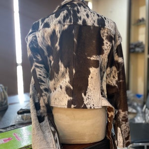 May include: A brown and white cowhide leather jacket with a zipper closure. The jacket has a cropped style and long sleeves.