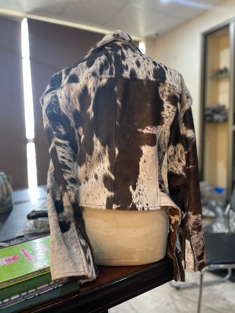 May include: A brown and white cowhide leather jacket with a zipper closure. The jacket has a cropped style and long sleeves.