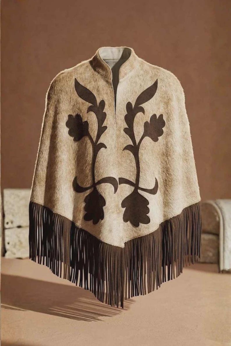 May include: A beige poncho with a textured, hair-on-hide appearance featuring a dark brown floral applique design on the front. The garment has a high collar and a long, dark brown fringe trim along the bottom hem, creating a pointed silhouette.