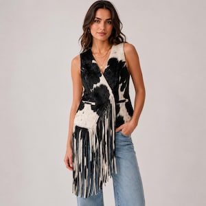 Op de afbeelding: Een mouwloos vest met een zwart-wit koeienprintpatroon, een V-hals en een lange asymmetrische zoom met franjes. Het kledingstuk heeft twee horizontale imitatiezakken aan de voorzijde en wordt gedragen met een lichtblauwe spijkerbroek.