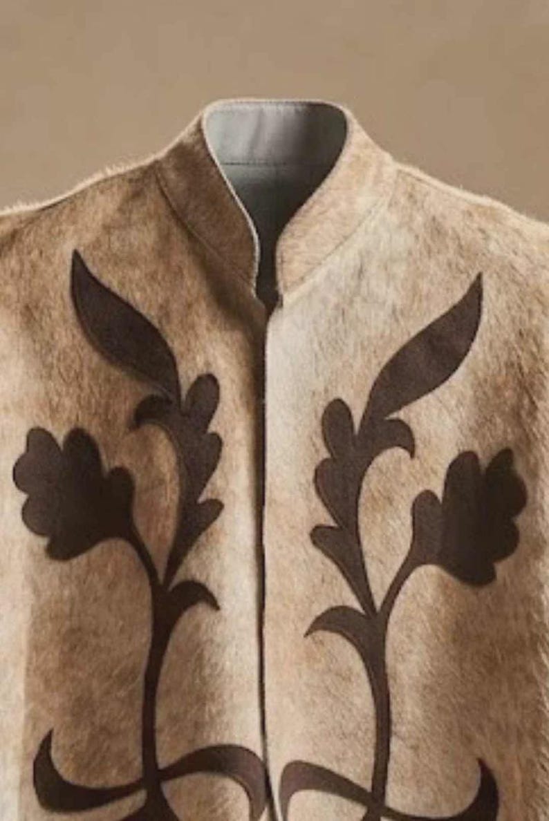 May include: A beige vest with a textured, fur-like appearance featuring a high collar and dark brown, leaf-shaped appliqu&eacute; designs on both sides of the front opening. The garment has a light gray interior lining visible at the neckline.