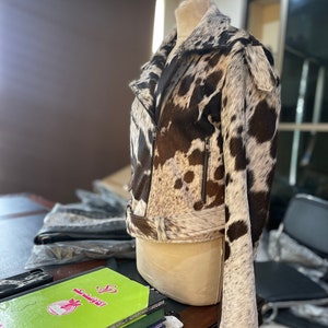 May include: A brown and white cowhide leather jacket with a zipper closure and a belt at the waist. The jacket is on a mannequin and is displayed in a room with a desk and chairs.