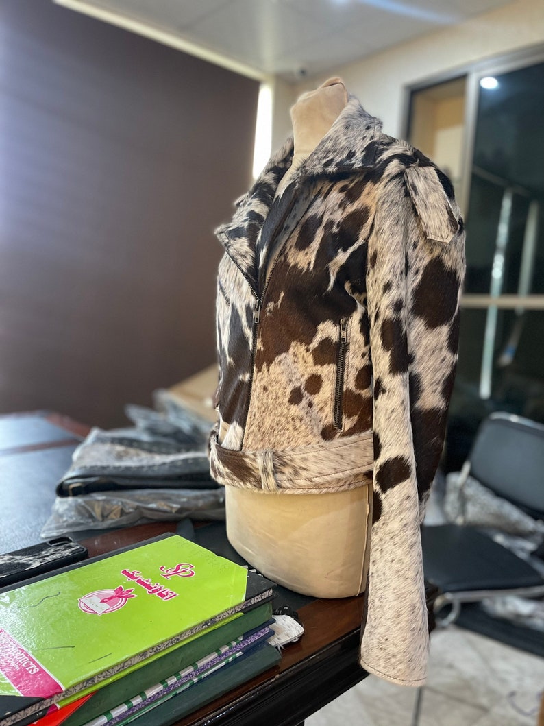 May include: A brown and white cowhide leather jacket with a zipper closure and a belt at the waist. The jacket is on a mannequin and is displayed in a room with a desk and chairs.