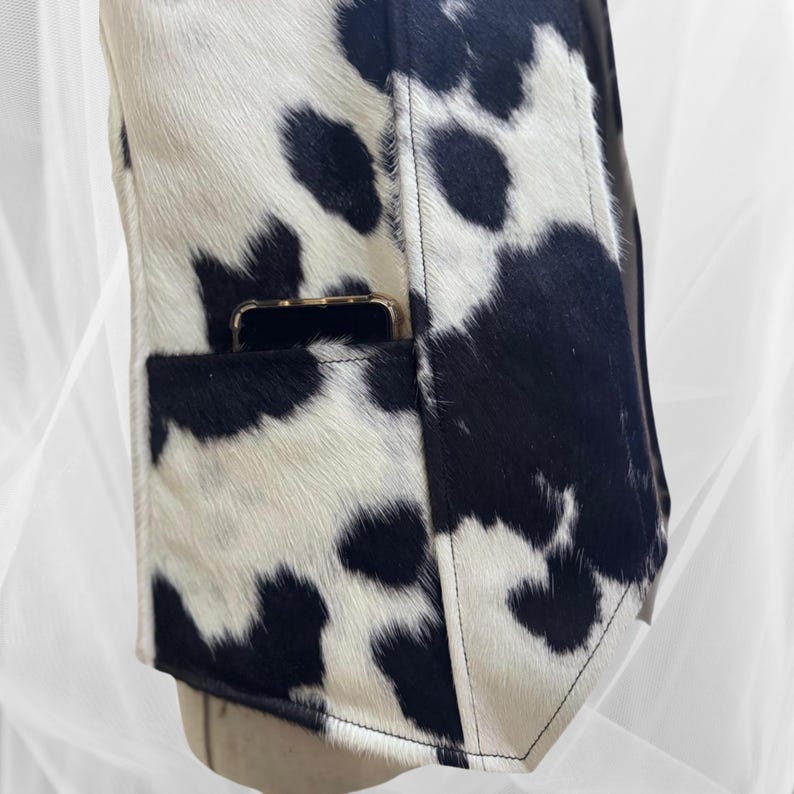 May include: A black and white cowhide jacket with a phone pocket on the left side. The jacket has a zipper closure and a collar.